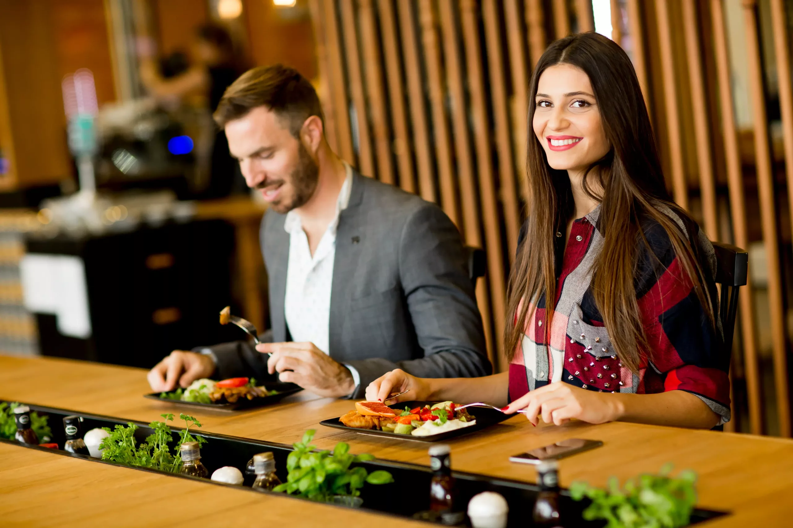 A man and a woman sit at a restaurant table in Abbotsford vs Chilliwack, enjoying salads. The woman smiles at the camera while the man looks down at his food.