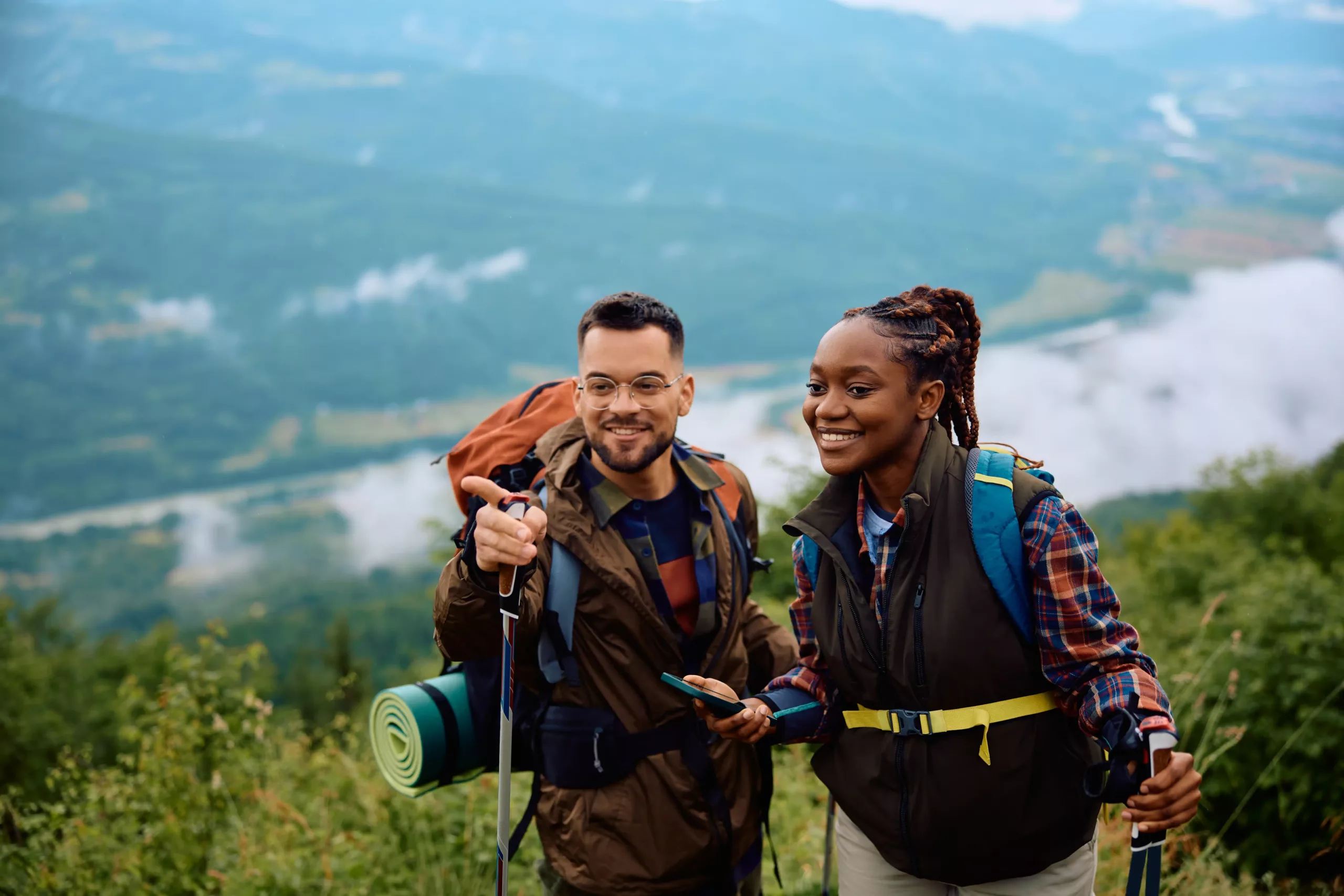 Two hikers with backpacks and trekking poles stand on a grassy hillside, smiling, enjoying views of a scenic valley and river—perfect for comparing Abbotsford vs Chilliwack outdoor adventures.