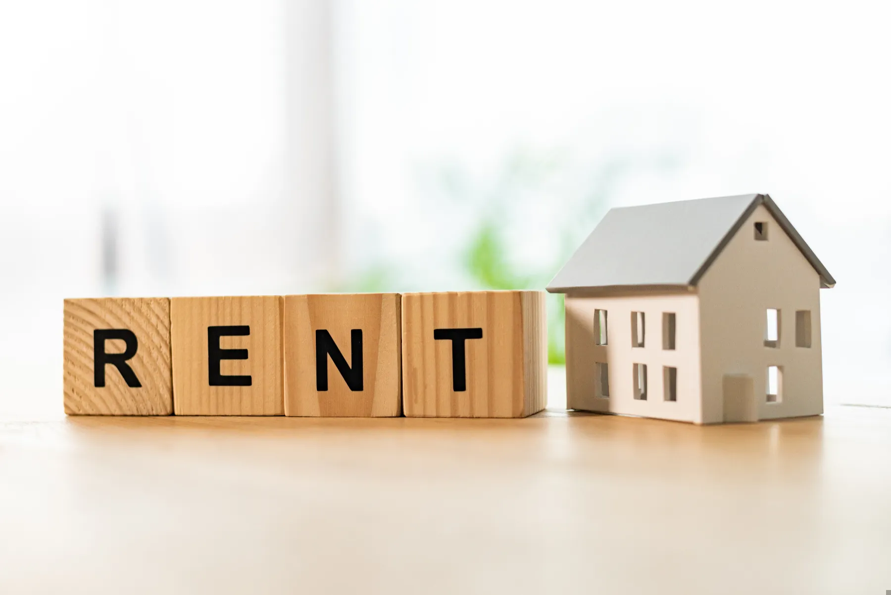 Wooden blocks spelling "RENT" are placed next to a small white model house on a light-colored surface with a blurred background.