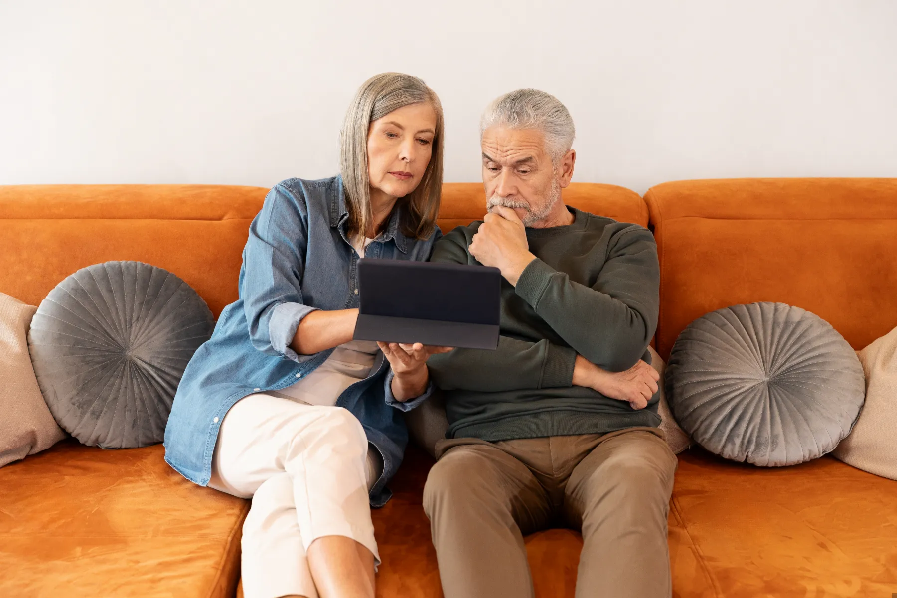 An older woman and man sit on an orange couch, looking intently at a tablet device together.