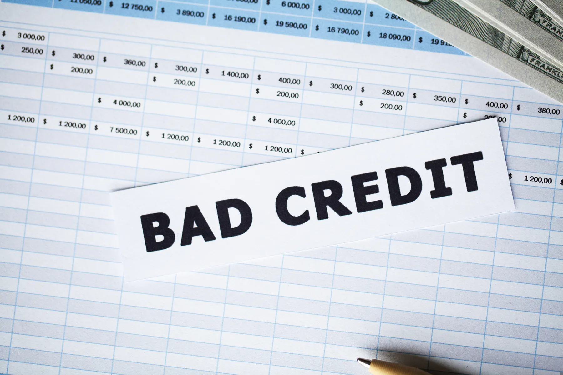 A piece of paper with "BAD CREDIT" written on it lies on top of a financial spreadsheet, with part of a pen and a stack of cash visible nearby.