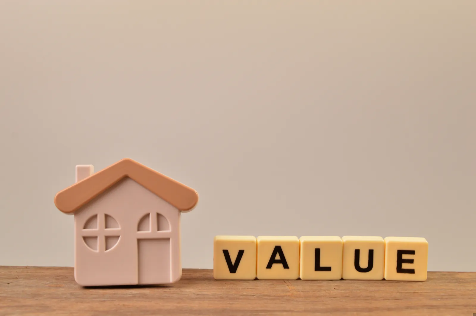 A small house model sits on a wooden surface next to letter tiles spelling "VALUE" against a plain background.