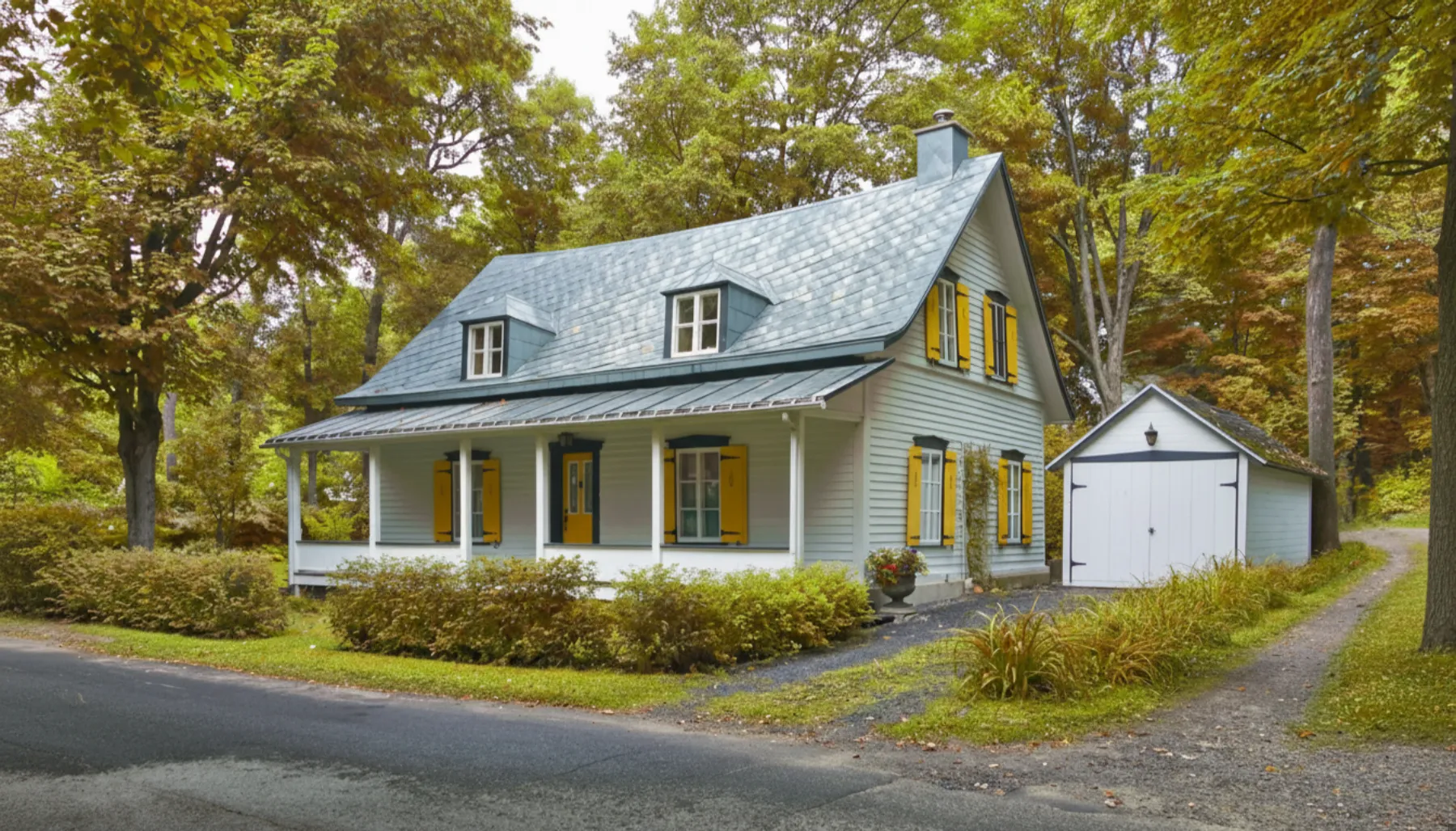 A light blue house with yellow shutters and a front porch sits next to a detached white garage, surrounded by trees and greenery.