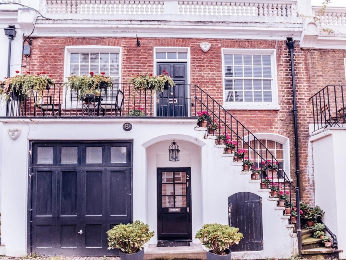 Red brick townhouse with black doors, white trim, and wrought iron stairs decorated with flowerpots. Upper level balcony has a small table, chairs, and potted plants.