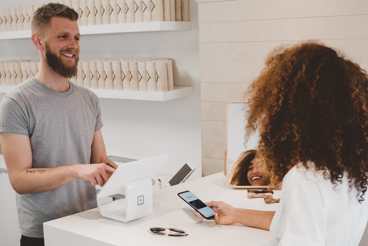 A man at a store counter smiles at a customer making a payment with her phone using a contactless card reader. Sunglasses and a mirror are on the counter.