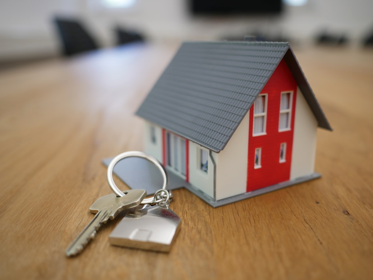 A small model house sits on a wooden table next to a set of keys with a house-shaped keychain.