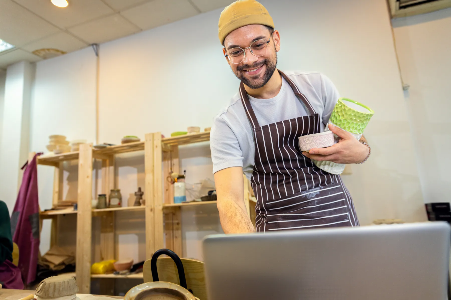 A man wearing an apron and beanie holds pottery while smiling and looking at a laptop in a ceramics studio with shelves in the background.