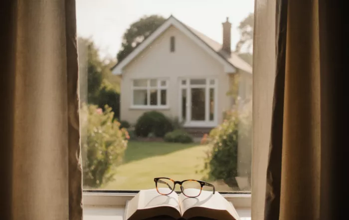 A pair of eyeglasses rests on an open book on a windowsill, with a blurred view of a house and garden outside the window.