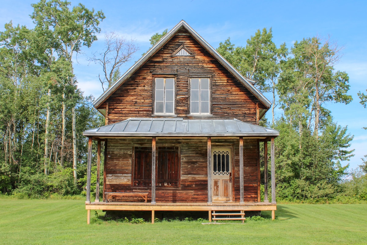 A weathered wooden house with a front porch, metal roof, and two upper windows, set on a grassy lawn with trees in the background under a clear sky.