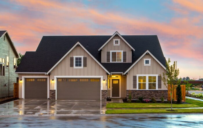 A two-story suburban house with three-car garage, light brown siding, stone accents, and a wet driveway at sunset.
