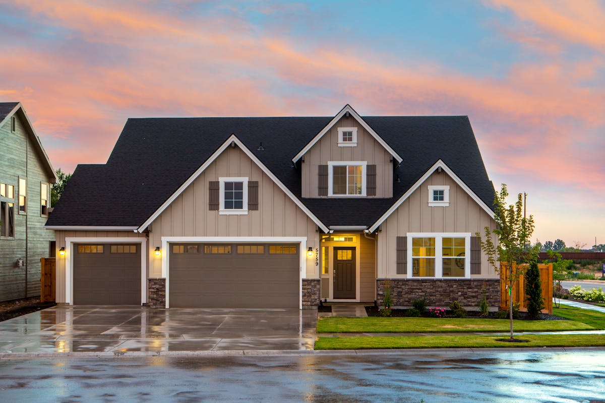 A two-story suburban house with three-car garage, light brown siding, stone accents, and a wet driveway at sunset.