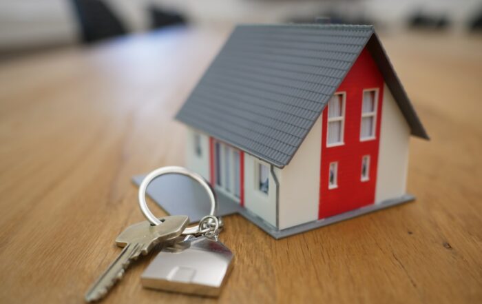 A small model house with a red door sits on a wooden table next to a set of keys on a keychain.