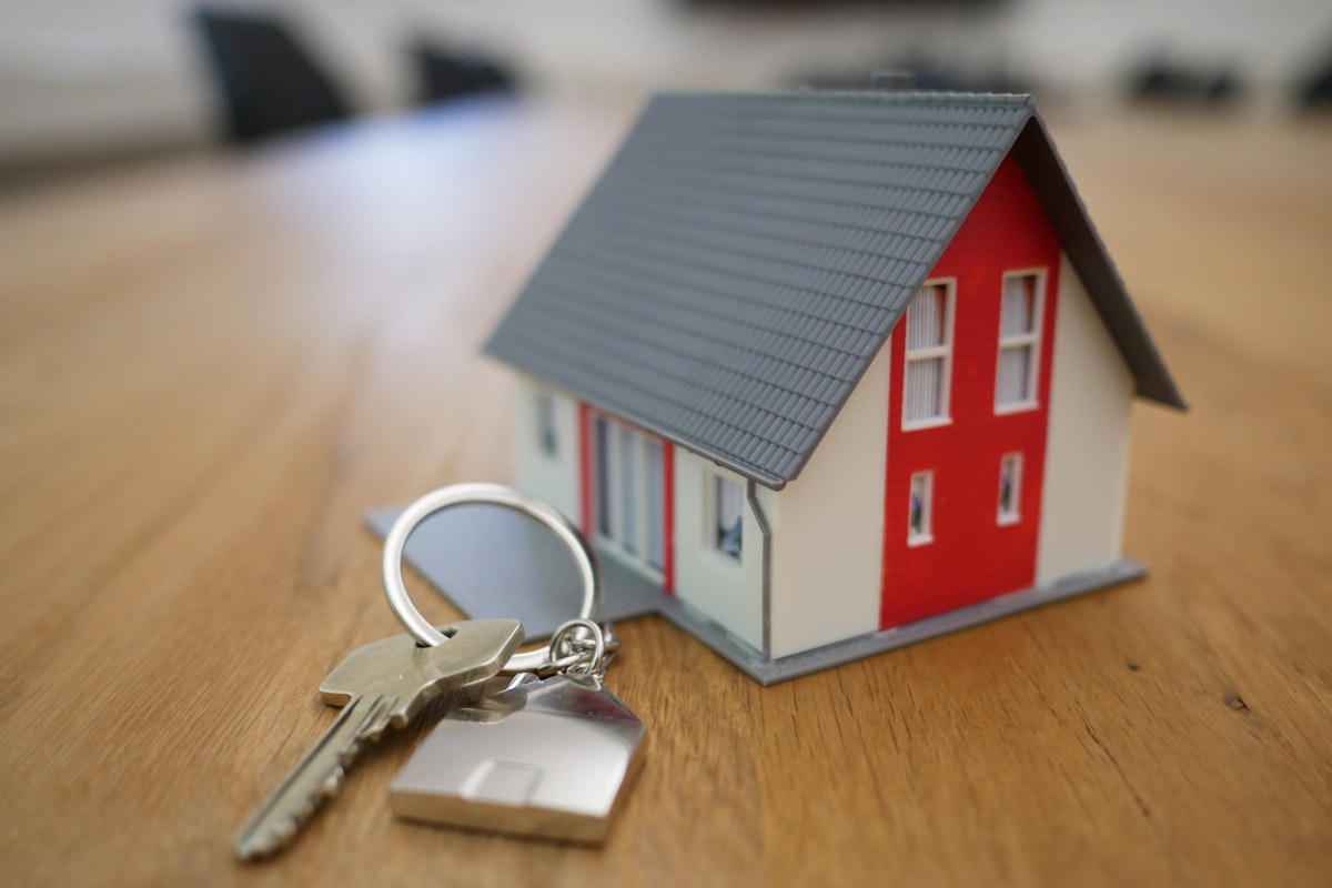 A small model house with a red door sits on a wooden table next to a set of keys on a keychain.